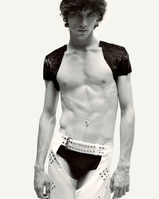 Male fashion model poses shirtless in white chaps with dark briefs against a neutral backdrop in a studio shot with moody lighting.