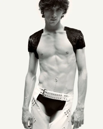 Male fashion model poses shirtless in white chaps with dark briefs against a neutral backdrop in a studio shot with moody lighting.