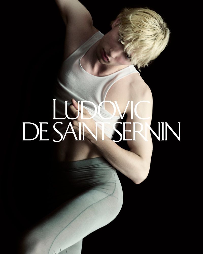 Male fashion model in a white tank top and grey leggings poses in a dramatic stance against a solid black backdrop in a studio.