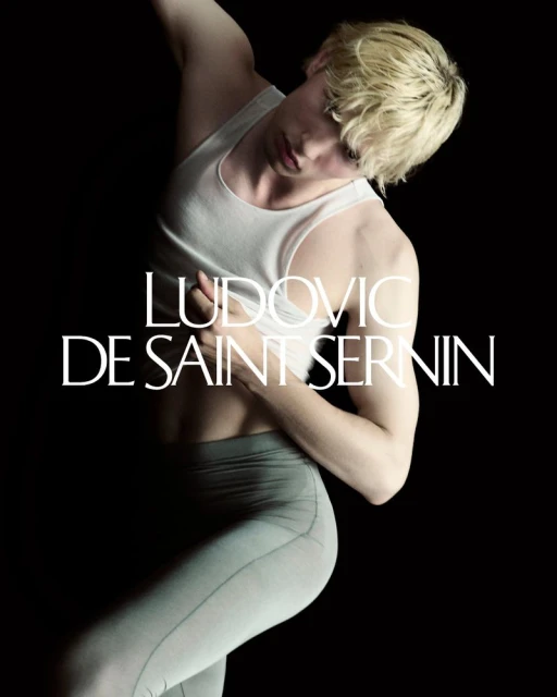 Male fashion model in a white tank top and grey leggings poses in a dramatic stance against a solid black backdrop in a studio.