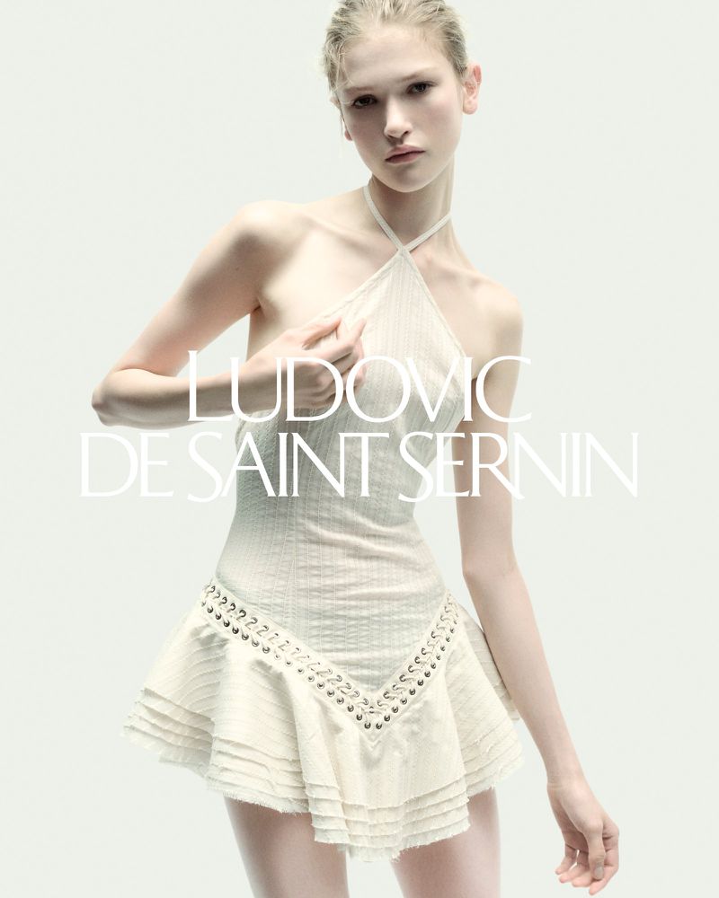 Fashion shot of a fair-skinned woman in a white halter dress with metal detailing, posing against a neutral background with visible text.