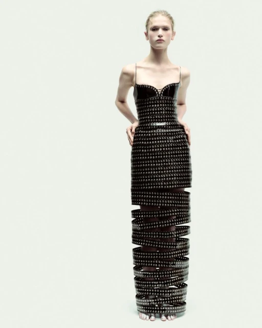 Elegant female model posing in a full-length black studded couture dress with openwork skirt, set against a minimalist white backdrop.