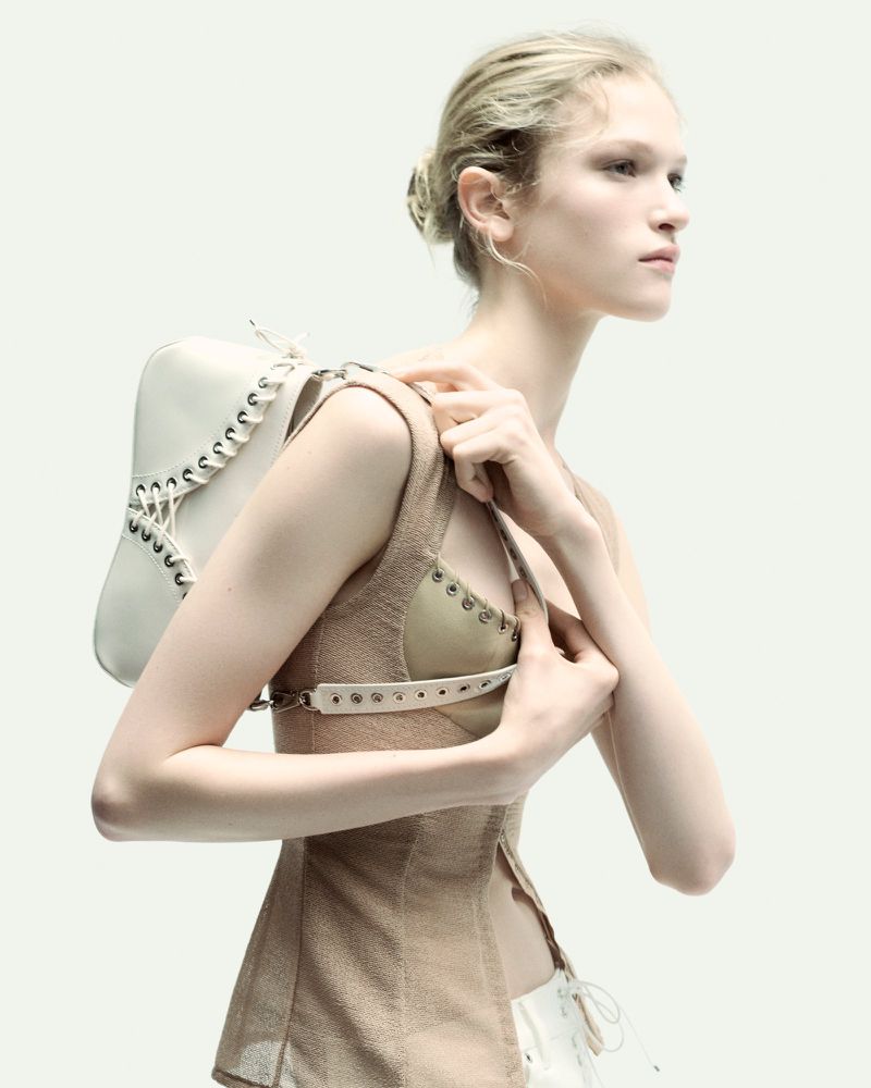 Blonde woman poses in a fashion shot wearing a beige top and white and beige bag with lacing against a bright white background.