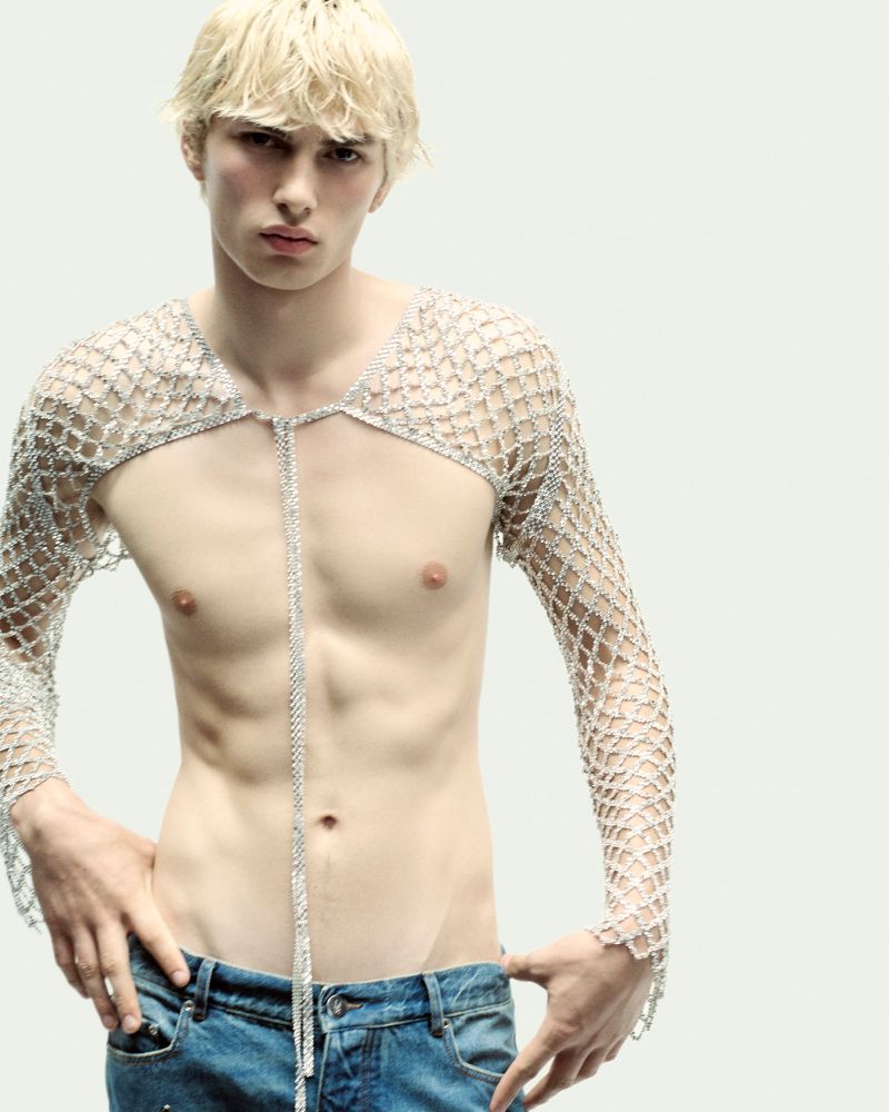 Male fashion model with blonde hair poses shirtless wearing a silver net top and blue jeans against a clean white background.