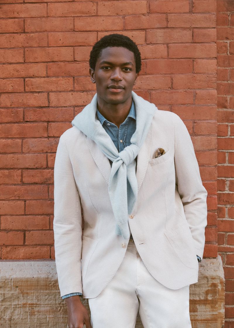 Black man in an off-white blazer and draped pale blue sweater posing in front of a red brick wall.