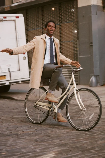A man wearing a trench coat rides a cream-colored bicycle on a cobblestone street, with a white delivery truck in the background.