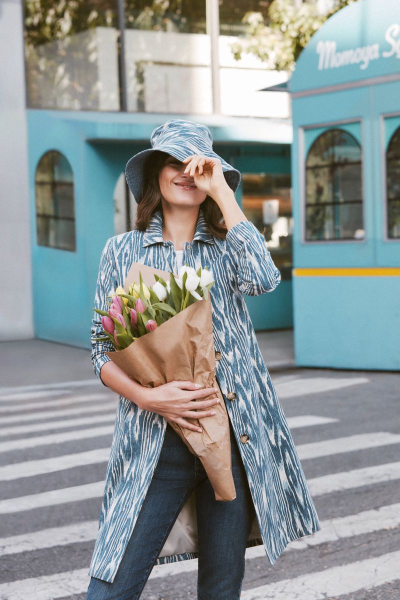 Adult woman stands outdoors on a crosswalk, holding a bouquet of flowers. She wears a patterned coat, bucket hat, and jeans.