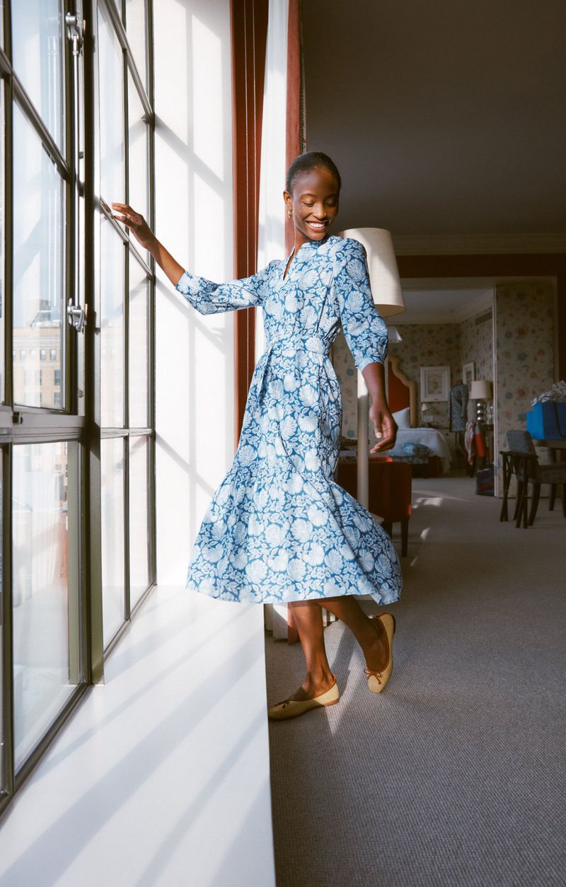 An elegant Black woman in a blue patterned dress stands by a window in a sunlit room. She wears flats and smiles in this fashion shot.