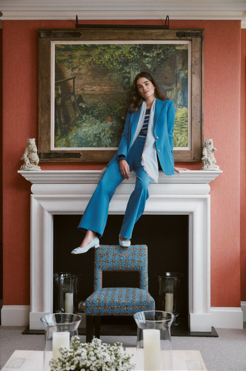 A stylish adult female in a blue pantsuit poses indoors on a chair beneath an oil painting, showcasing a blend of fashion and interior design.