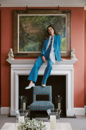 A stylish adult female in a blue pantsuit poses indoors on a chair beneath an oil painting, showcasing a blend of fashion and interior design.