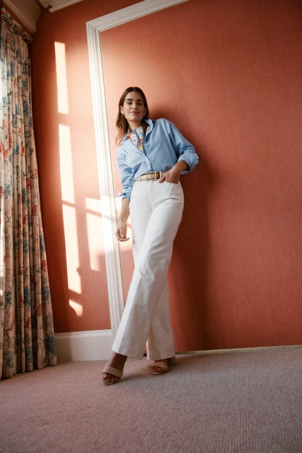 A stylish adult woman poses indoors wearing a light blue shirt, white jeans, and nude heels, standing near a peach-colored wall and patterned curtain.