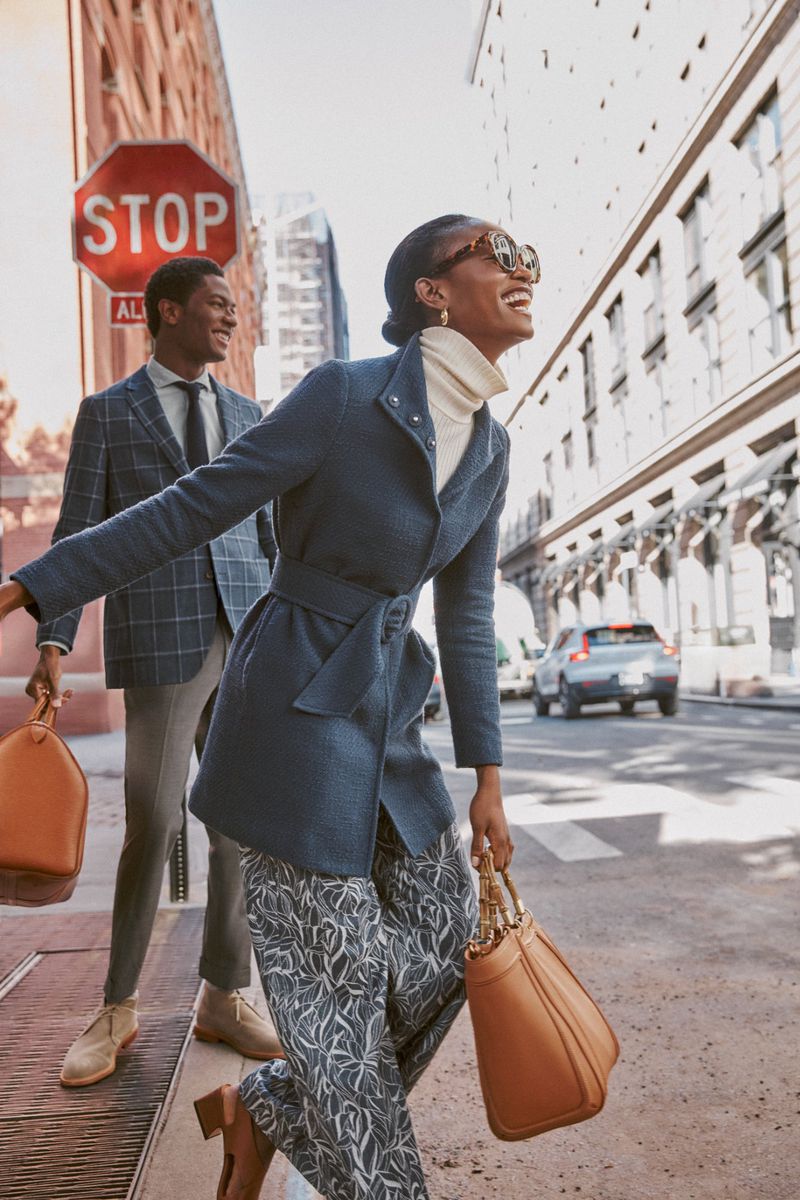 Smiling woman in blue belted coat and patterned pants walking on city street holding a brown leather bag with a man behind her