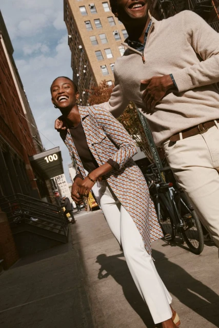 Two smiling people, a man and a woman, walking on a city sidewalk with buildings in the background on a sunny day, medium shot.