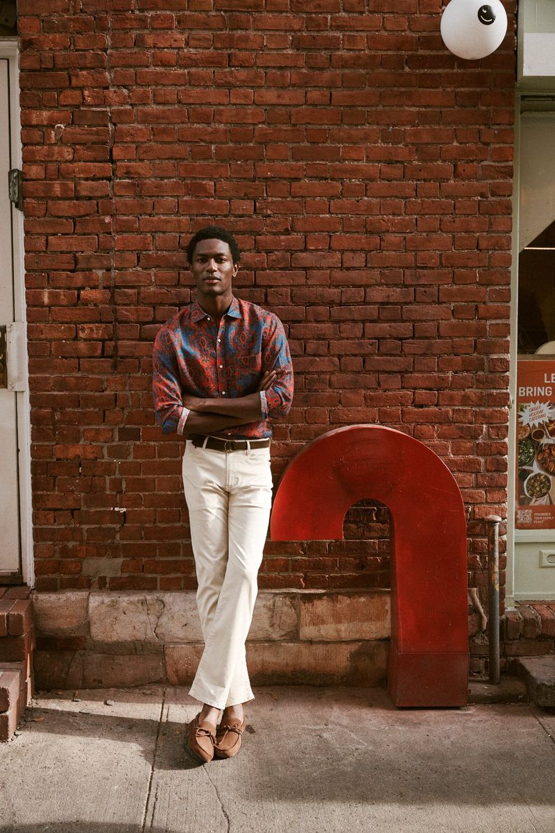 Man in patterned shirt and white pants stands next to a red, curved, metal sculpture in front of a brick building, looking at camera.