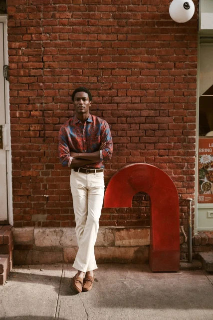 Man in patterned shirt and white pants stands next to a red, curved, metal sculpture in front of a brick building, looking at camera.