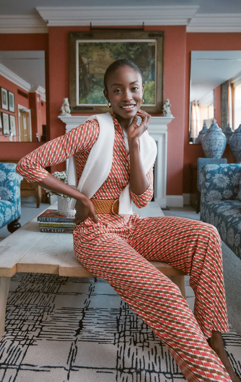 Indoor portrait of a smiling Black woman in a patterned jumpsuit sitting on a coffee table with decor in the background.