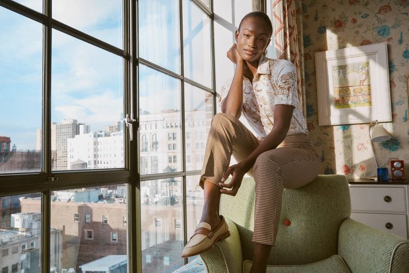 A dark-skinned woman sits indoors in neutral patterned clothing, posing on a green armchair, lit by daylight from a large window.