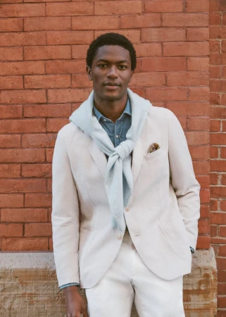 Black man in an off-white blazer and draped pale blue sweater posing in front of a red brick wall.