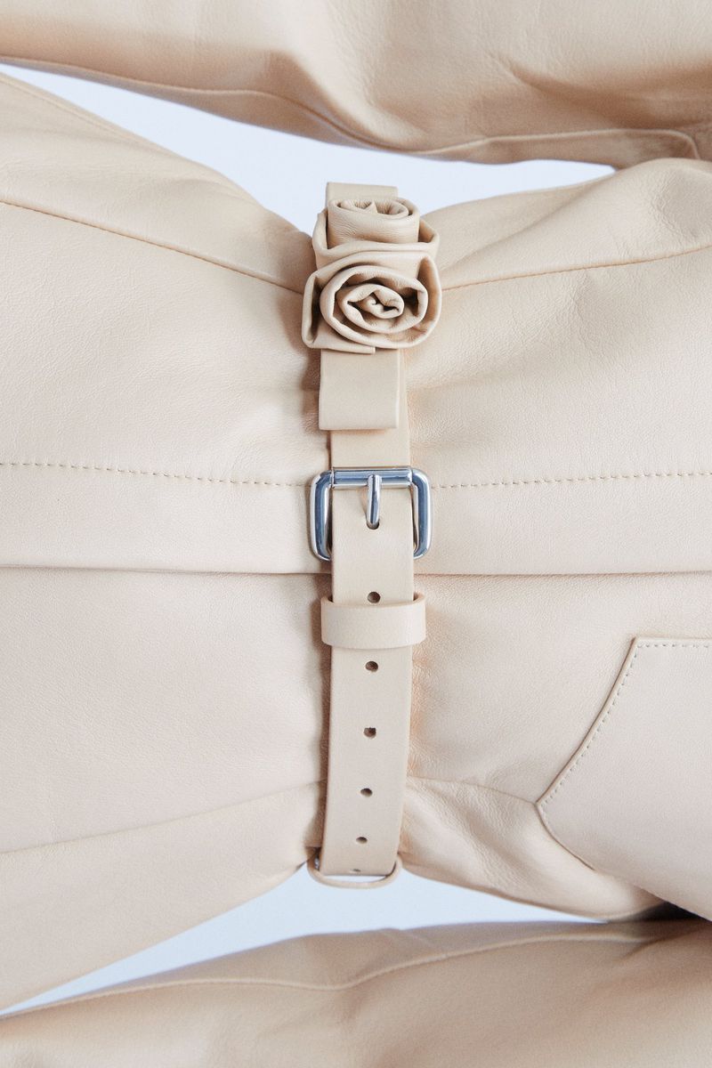 Close-up of a light beige leather garment with a decorative rose and buckle strap accent, set against a bright white background.