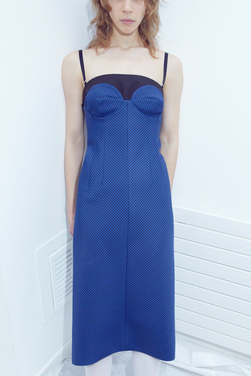 Model standing against a plain white interior wall wearing a blue patterned midi dress with black straps.