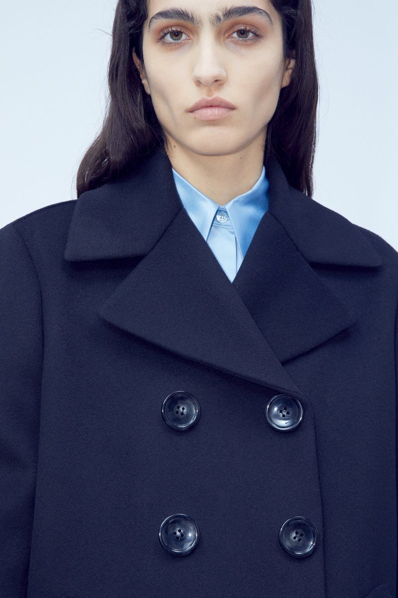 Woman in a navy overcoat and a light blue collared shirt posing against a white backdrop in a studio setting, fashion advertisement.