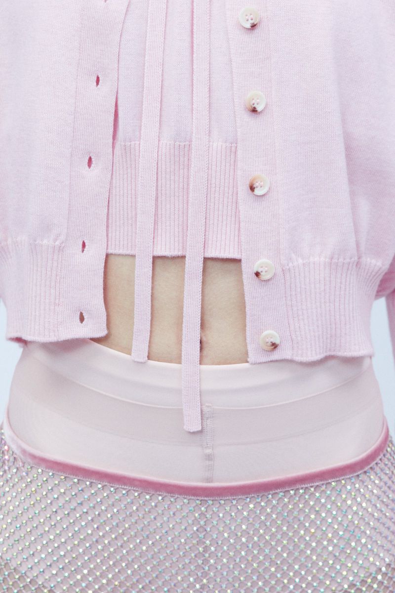 Close-up of a woman's midriff wearing a pink knit cardigan, top with strings, and a sparkling silver skirt in a light, indoor setting.
