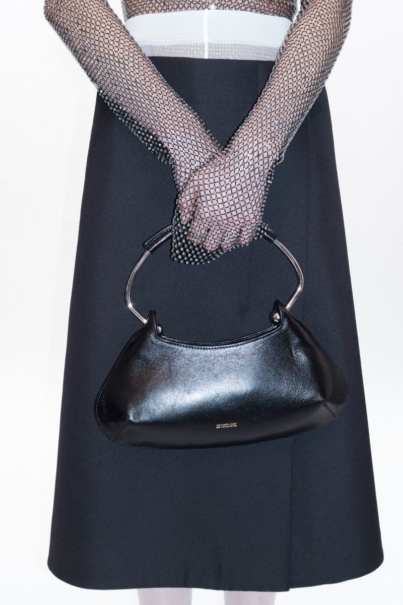 Close-up fashion shot of a woman holding a black handbag with silver handles, wearing black gloves and a black skirt on a white background.