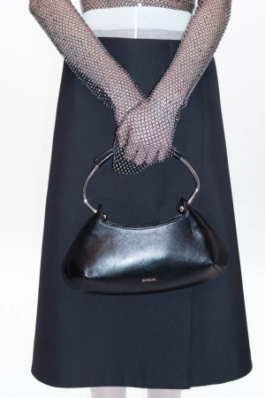 Close-up fashion shot of a woman holding a black handbag with silver handles, wearing black gloves and a black skirt on a white background.