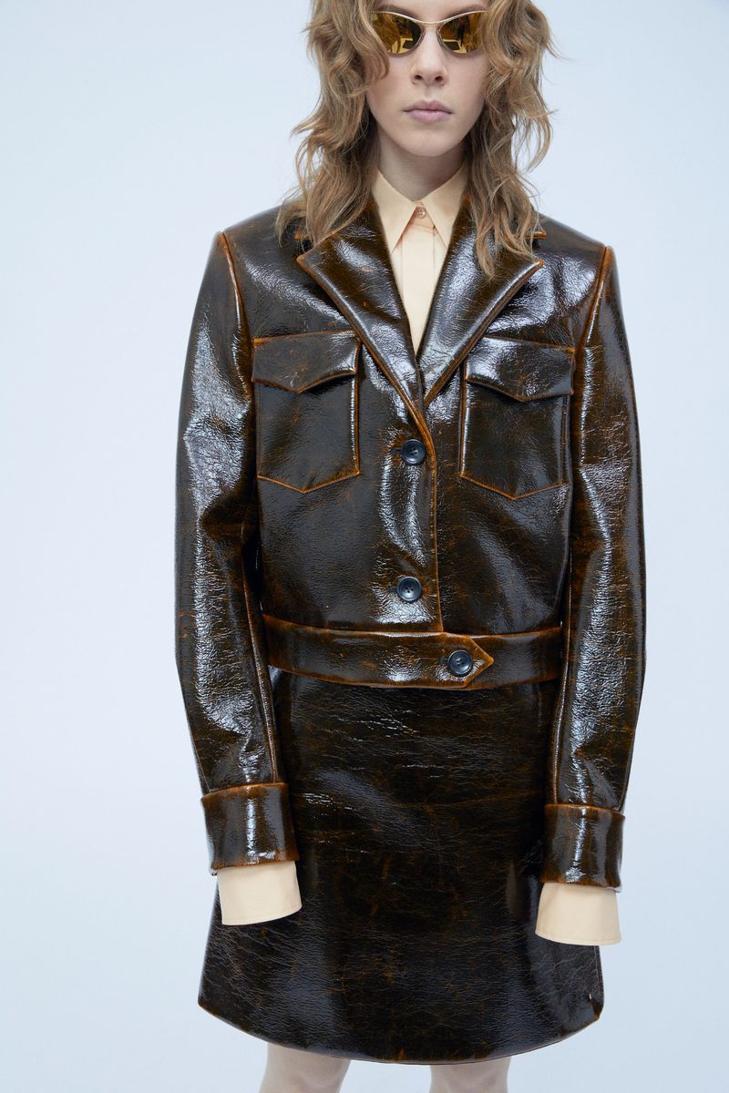 A fair-skinned model presents a shiny brown jacket and skirt ensemble with orange trim against a bright white backdrop in a fashion shot.