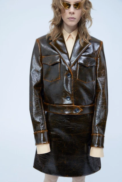 A fair-skinned model presents a shiny brown jacket and skirt ensemble with orange trim against a bright white backdrop in a fashion shot.