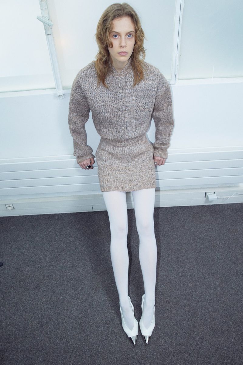 Model wearing a textured knit top, mini skirt, and white tights against a white wall.