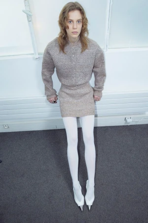 Model wearing a textured knit top, mini skirt, and white tights against a white wall.