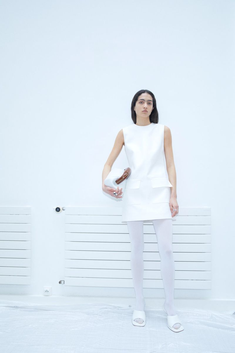 A slender woman in a white mini dress and sheer tights, holding a handbag, stands against a white wall and radiator in a studio.