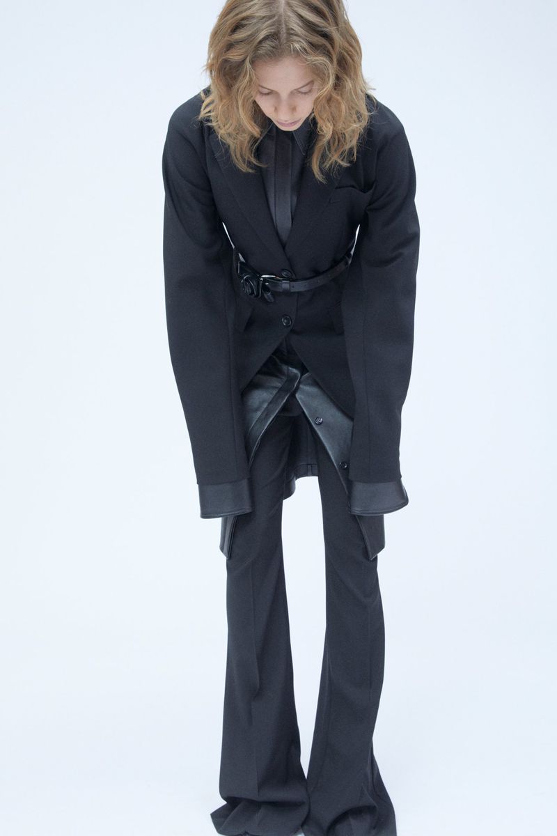 Model in black suit with oversized blazer and flared trousers posing against white seamless background