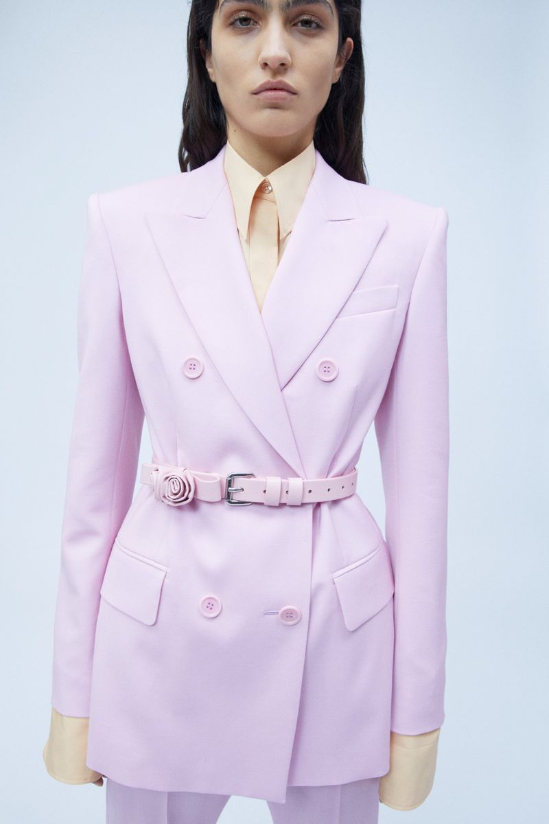 Fashionable woman in a pink suit. Model poses in a double-breasted pink blazer with a matching belt and trousers against a plain background.