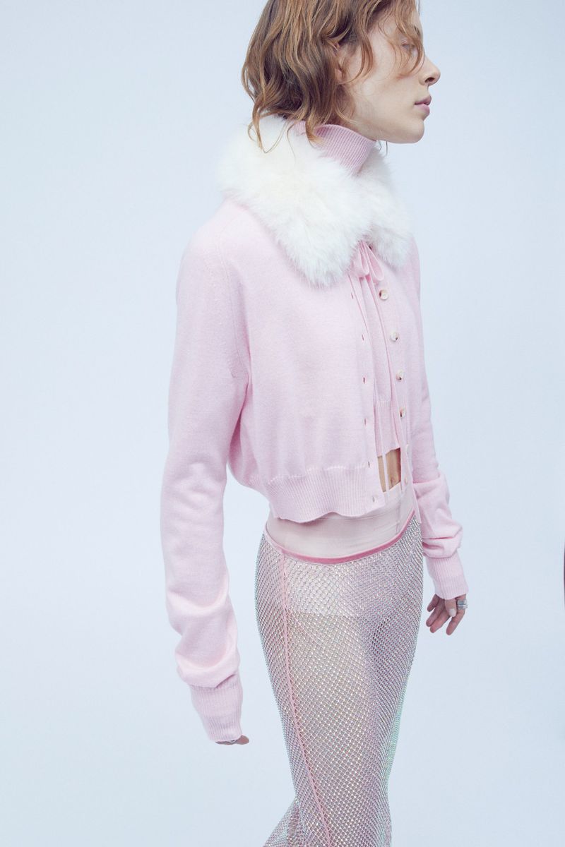 Woman in pink cardigan with fur collar and shimmering pants poses in a studio with soft, diffused lighting against a light blue background.