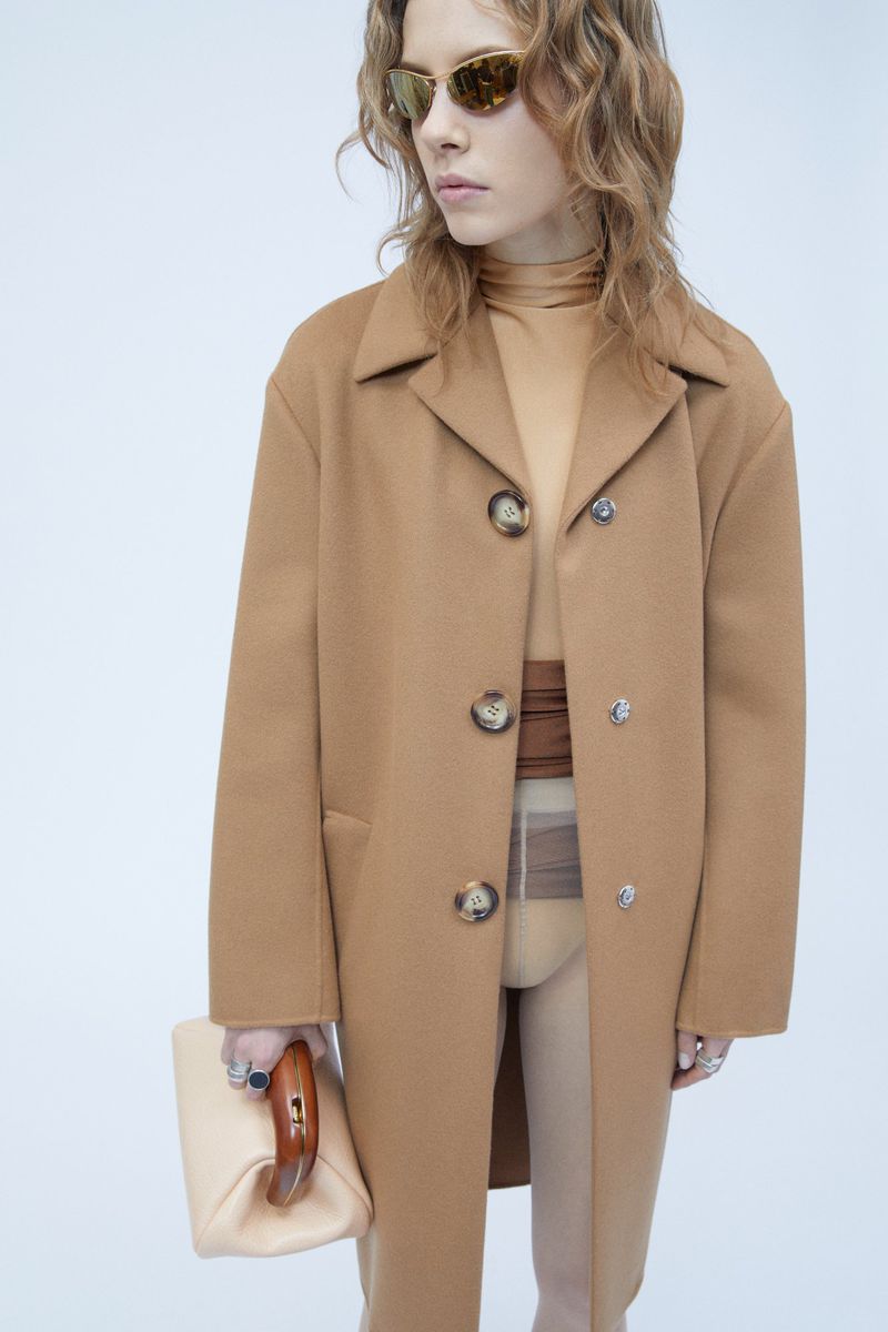 Model wearing a camel coat and sunglasses holding a light handbag against a white background