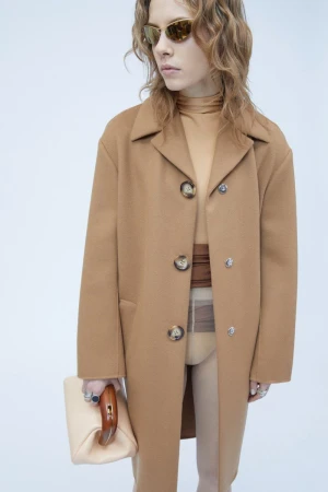 Model wearing a camel coat and sunglasses holding a light handbag against a white background