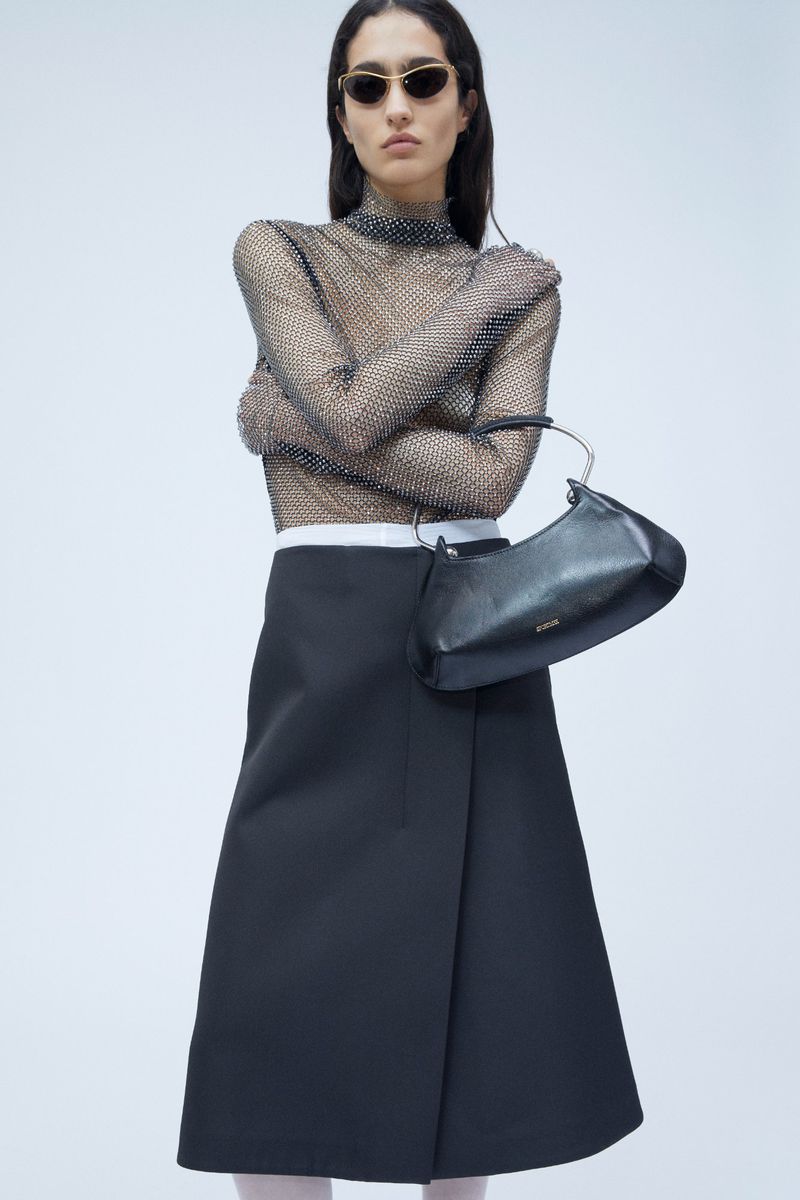 A woman in a sheer, embellished top and black skirt poses with a black handbag against a plain backdrop in a fashion studio shot.