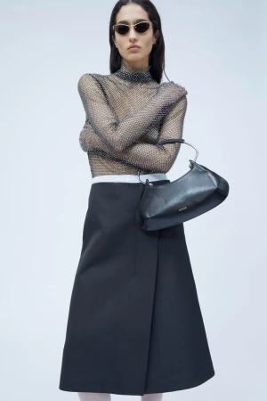 A woman in a sheer, embellished top and black skirt poses with a black handbag against a plain backdrop in a fashion studio shot.