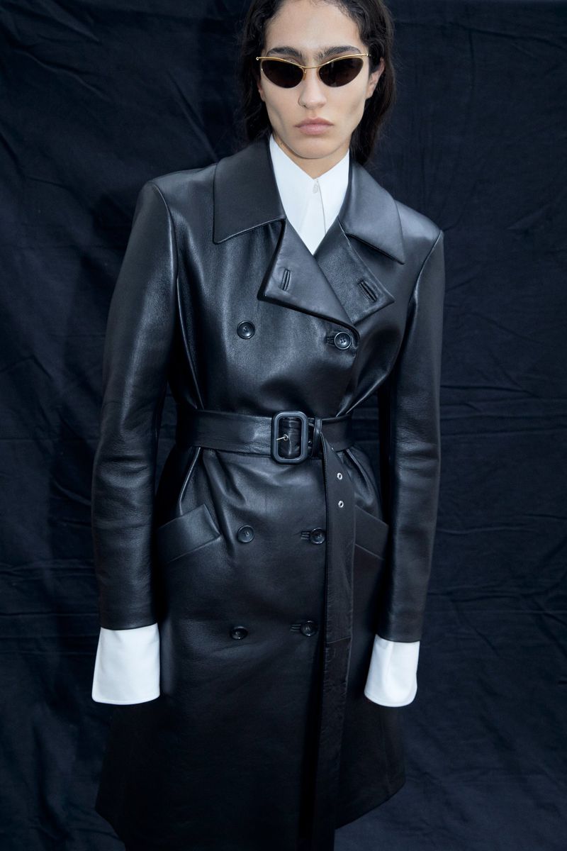 A woman poses in a black leather trench coat and sunglasses against a dark backdrop, showcasing a blend of high fashion and edgy style.