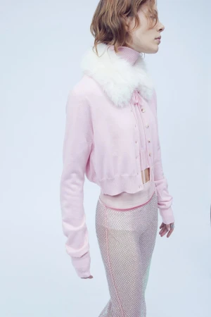 Model wearing a pink cardigan with white faux fur collar and textured sheer pants against a light background
