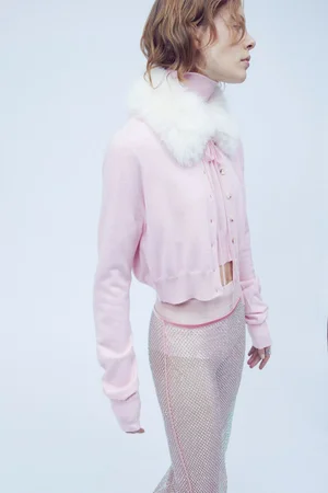 Model wearing a pink cardigan with white faux fur collar and textured sheer pants against a light background