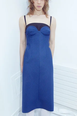 Model standing against a plain white interior wall wearing a blue patterned midi dress with black straps.