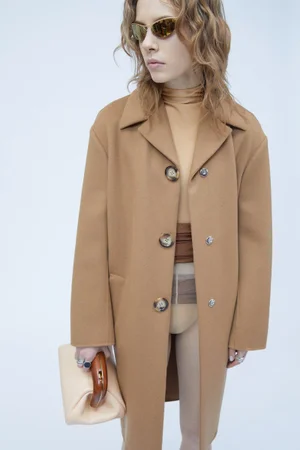 Model wearing a camel coat and sunglasses holding a light handbag against a white background