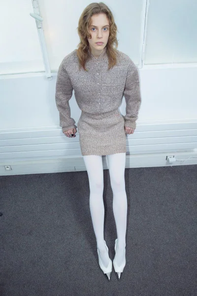 Model wearing a textured knit top, mini skirt, and white tights against a white wall.