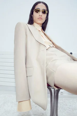 Model in cream blazer and sunglasses seated on a stool against a plain background for Sportmax Pre Fall 2024 lookbook.
