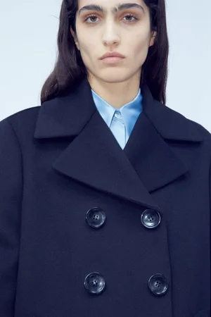 Model wearing a dark navy double-breasted coat over a light blue collared shirt against a pale studio backdrop.