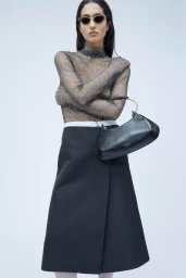 Model wearing dark sunglasses, sheer mesh top, and black midi skirt holding a black leather shoulder bag against a white backdrop.