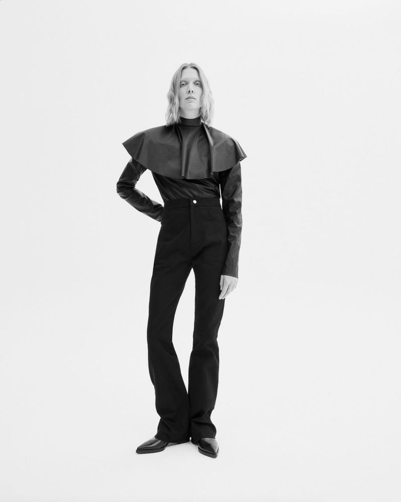 Fashion portrait of a model in black high-waisted pants, long-sleeved black top, and black pointy shoes against a white backdrop.